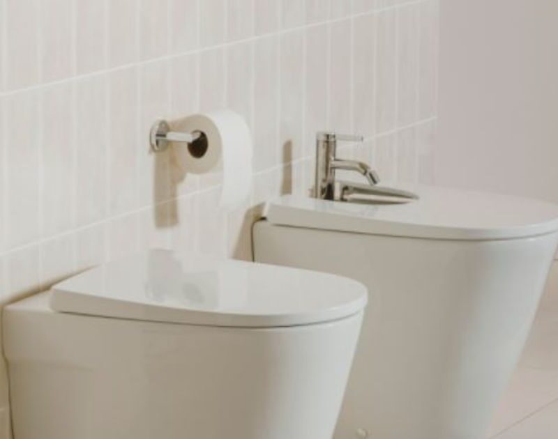 baños Baño minimalista con inodoro y bidé blancos, paredes de azulejos rosados suaves y una cesta de mimbre, destacando un diseño limpio y sereno